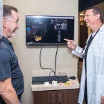 Dr. Stephen Wilson reviewing 3D digital imaging scans on a wall monitor with a male patient.
